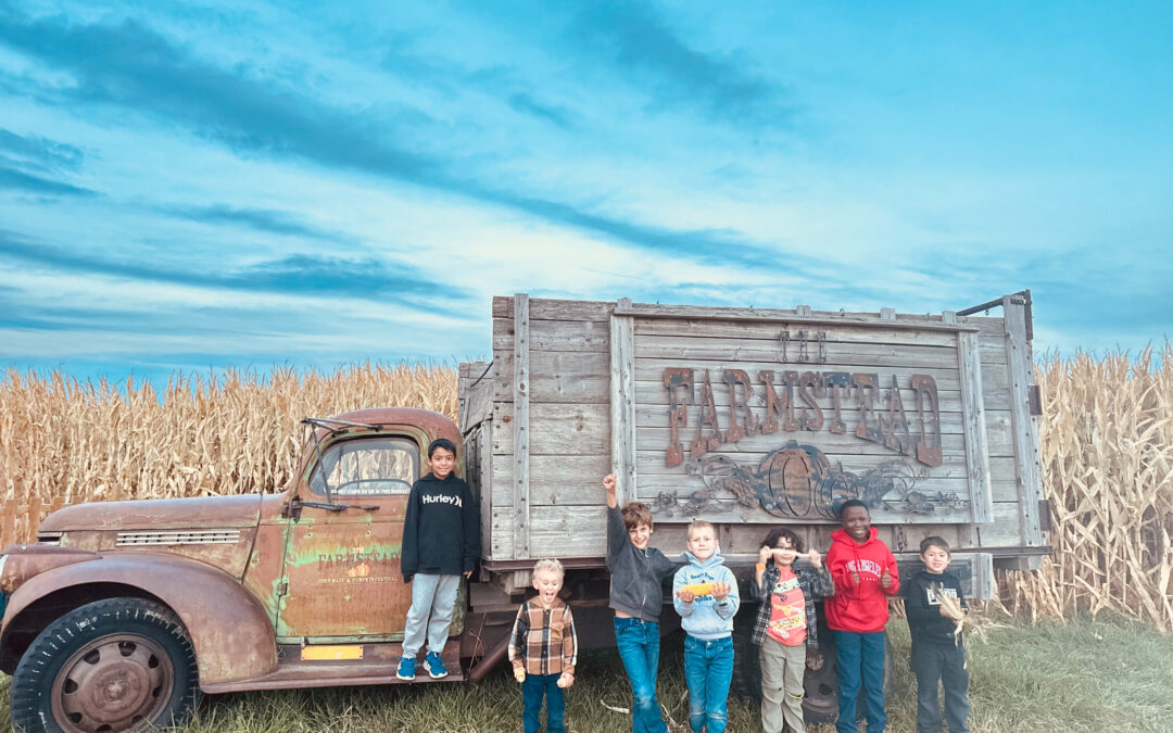 Kids lined up beside a vintage Farmstead farm truck in front of a tall corn maze on a fall afternoon