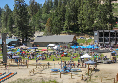 Bogus Basin base area in summer with tents, lawn seating, a climbing wall and bungee trampolines below the pine-covered mountain